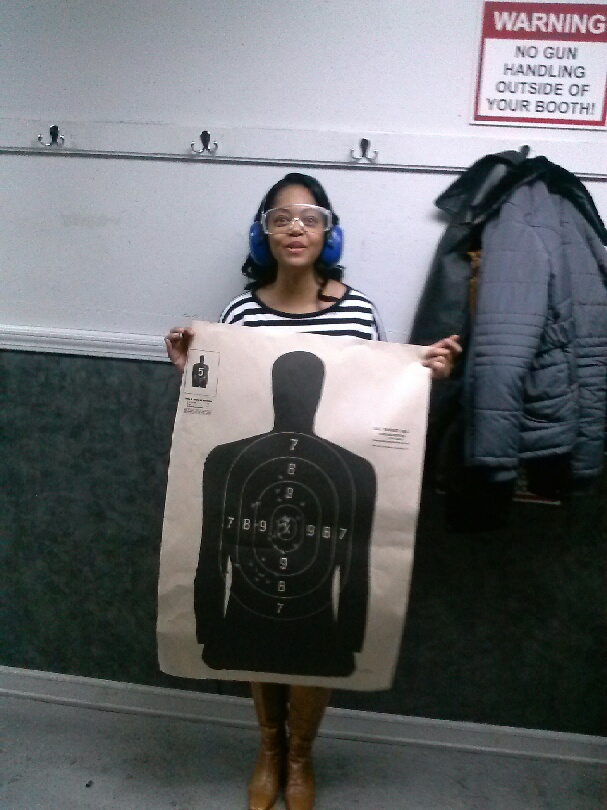 Karen Whitsett wearing eye and ear protection at an indoor shooting range, holding a paper silhouette target.